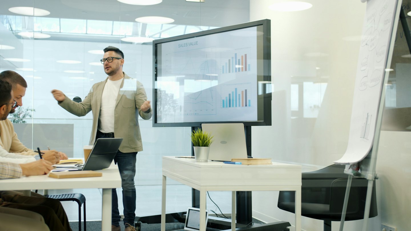 Presenter discusses sales charts with colleagues in a modern office.